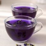Clear glass cup filled with vibrant purple Brazilian Mounjaro drink made from butterfly pea flower, on a bright marble surface