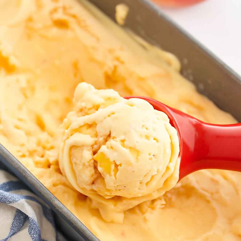 A close-up view of a scoop of peach frozen yogurt with visible peach chunks, lifted from a loaf pan with a teal-handled scoop.