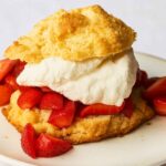 Close-up of a strawberry shortcake with golden biscuit, whipped cream, and fresh sliced strawberries on a white plate.