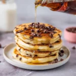 Fluffy chocolate chip pancakes stacked on a plate with syrup drizzle