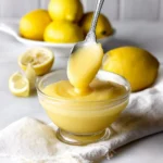 A jar of homemade lemon curd surrounded by fresh lemons and a spoon