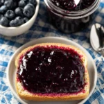 Homemade blueberry jam in a glass jar with blueberries and bread on the side